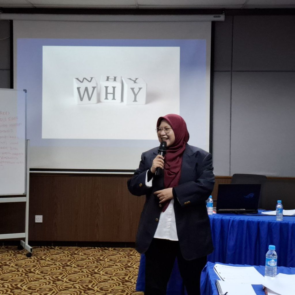 Jurulatih HRDC bertudung merah dan blazer hitam sedang menyampaikan latihan korporat.