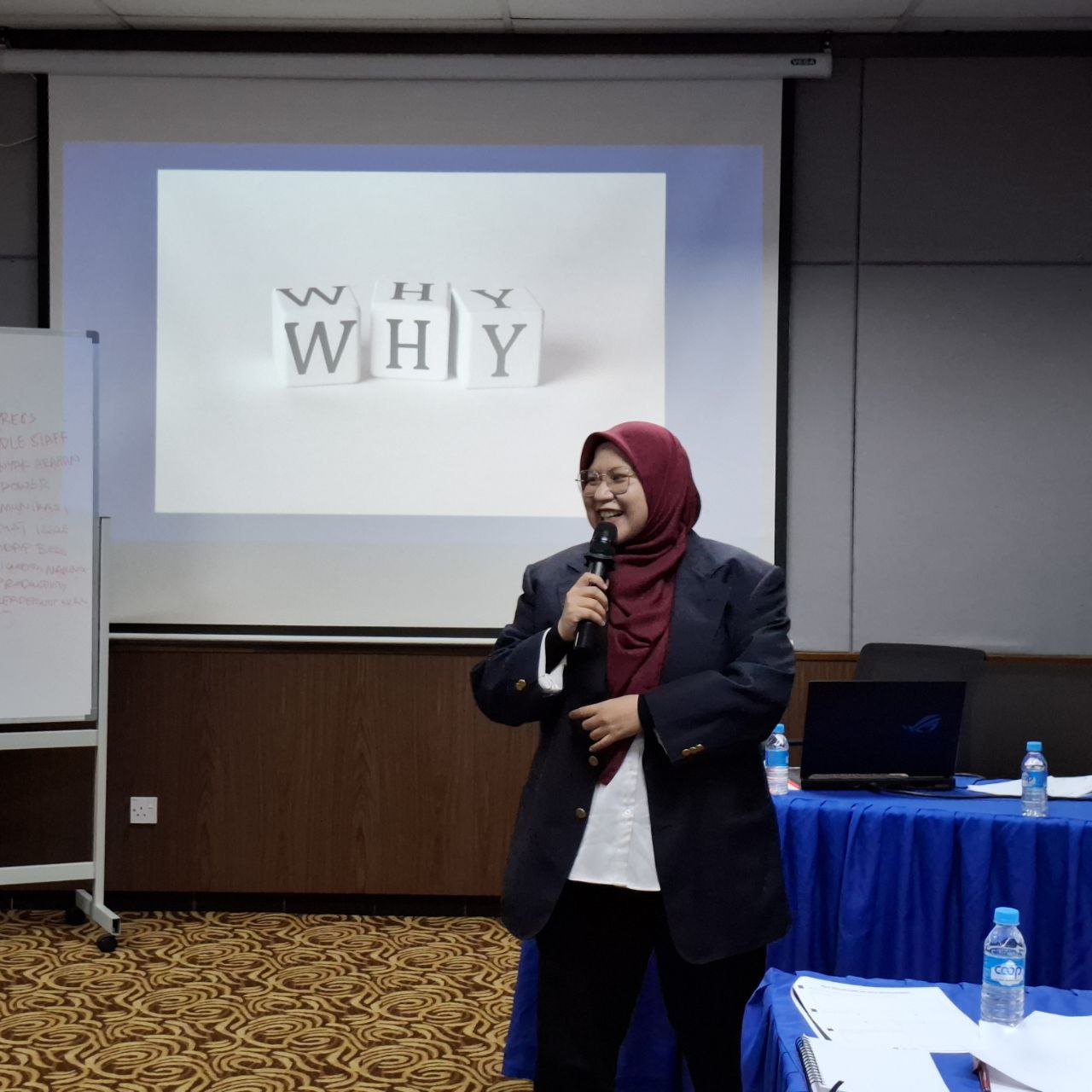 Jurulatih HRDC bertudung merah dan blazer hitam sedang menyampaikan latihan korporat.