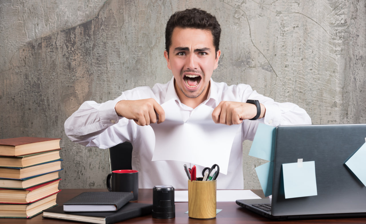 Pekerja pejabat marah mengoyakkan kertas di meja dengan laptop dan buku, melambangkan stres kerja dan tekanan emosi.