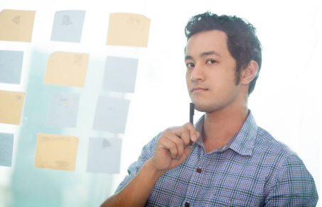 Young professional in checkered shirt holding a pen and thinking, with sticky notes on glass wall in the background, brainstorming video content ideas