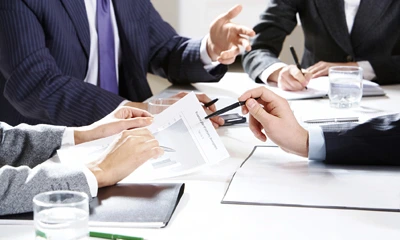 Pandangan dekat sekumpulan ahli profesional dalam mesyuarat perniagaan, sedang menganalisis dokumen dan carta data di atas meja putih.