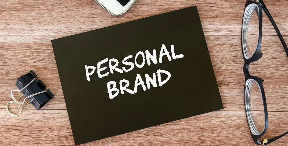 Kad hitam dengan perkataan putih tebal "PERSONAL BRAND" diletakkan di atas permukaan meja kayu. Terdapat sepasang cermin mata berbingkai hitam dan pengapit kertas hitam di sekeliling kad tersebut, menonjolkan fokus kepada penjenamaan peribadi yang profesional dan konsisten.