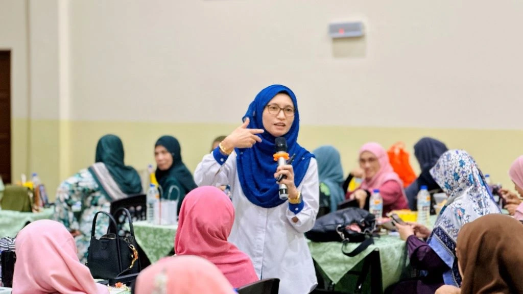 Seorang penceramah wanita yang memakai tudung biru sedang memberikan ucapan sambil memegang mikrofon kepada sekumpulan wanita yang menjadi audiens. Ekspresi wajah penceramah melambangkan ketenangan dan keyakinan, yang menekankan kepada konsep Kepimpinan Wanita yang berlandaskan Ketenangan dan Nilai Murni dalam persekitaran seminar.