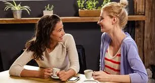 Dua orang wanita sedang duduk berbincang sambil tersenyum di sebuah kafe dengan cawan kopi di atas meja, menggambarkan suasana sokongan emosi dan perkongsian pengalaman secara santai namun bermakna di antara rakan sekerja.