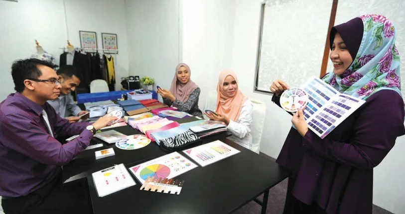 Sekumpulan wanita berkerjaya yang memakai tudung sedang berbincang dengan rakan sekerja lelaki di sebuah meja mesyuarat sambil meneliti katalog warna pakaian, mencerminkan penampilan diri yang kemas, profesional, dan ceria di tempat kerja.