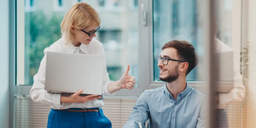 Seorang wanita sedang berdiri sambil memegang komputer riba dan memberikan isyarat "ibu jari bagus" (thumbs up) kepada seorang rakan sekerja lelaki yang sedang duduk, melambangkan pemberian maklum balas positif dan pengiktirafan di tempat kerja.