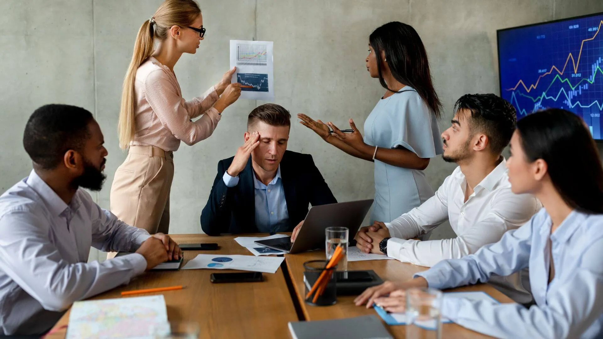 Suasana mesyuarat yang tegang di mana seorang pemimpin lelaki kelihatan stres sambil memegang dahi, manakala rakan sekerja wanita di sisinya menunjukkan dokumen dengan bahasa tubuh yang agresif, menggambarkan kepentingan mengawal ekspresi wajah dan emosi untuk mengekalkan profesionalisme.
